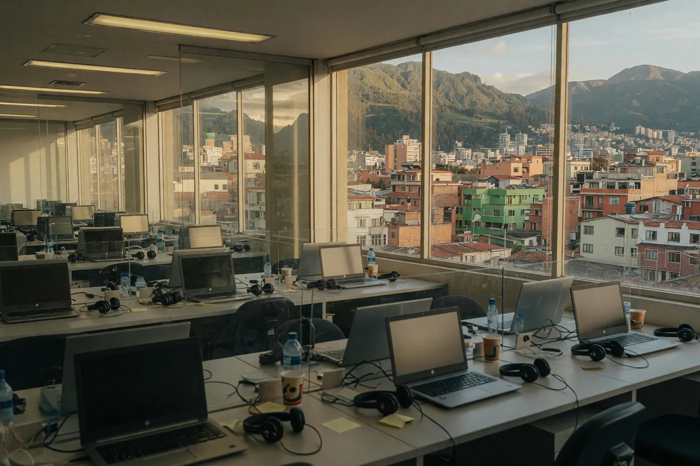 Rows of customer-support workstations with headsets and laptops in a Colombian contact center office, city towers visible through the windows