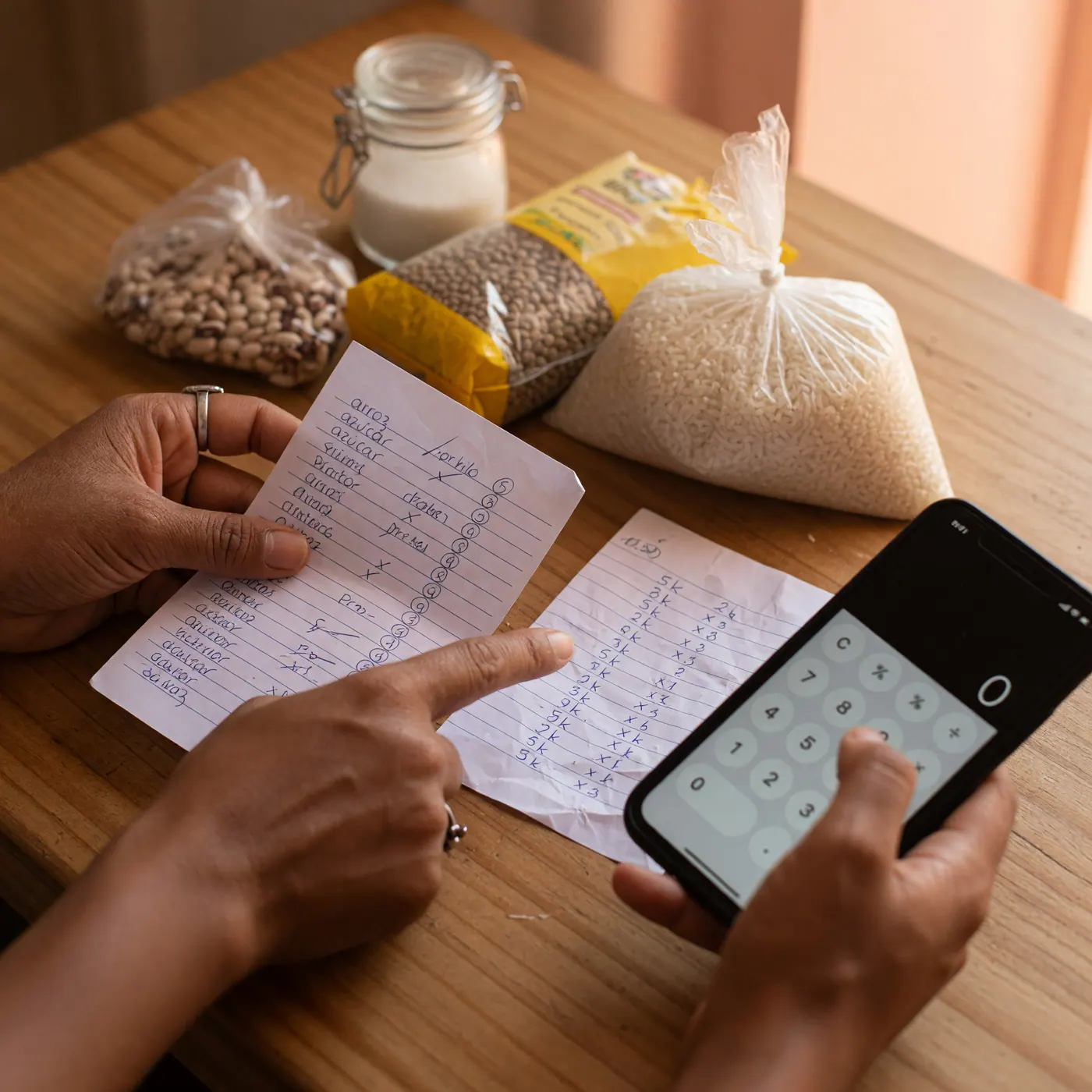 Hands comparing unit prices on a handwritten grocery list beside pantry staples and a phone calculator