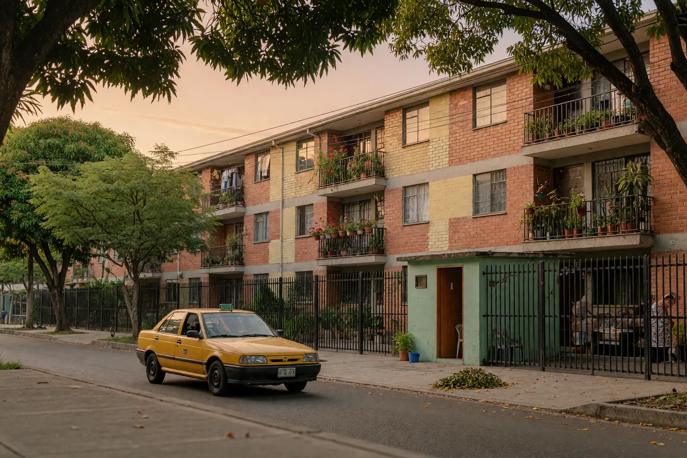 A quiet, tree-lined residential street in Colombia with a low-rise red-brick apartment building, security entrance, green balconies, and a yellow taxi driving past.
