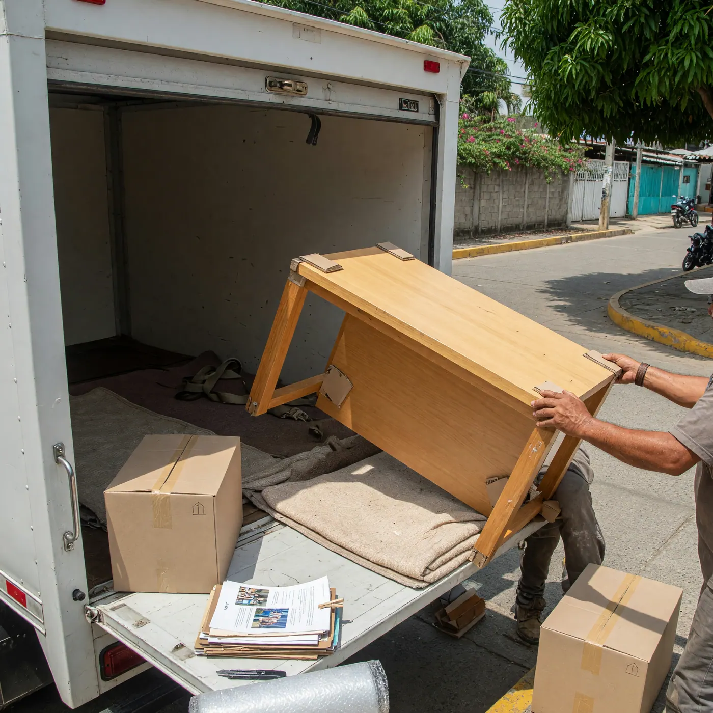 Un transportador carga una mesa de madera y cajas en un camión blanco de mudanzas en una calle residencial.
