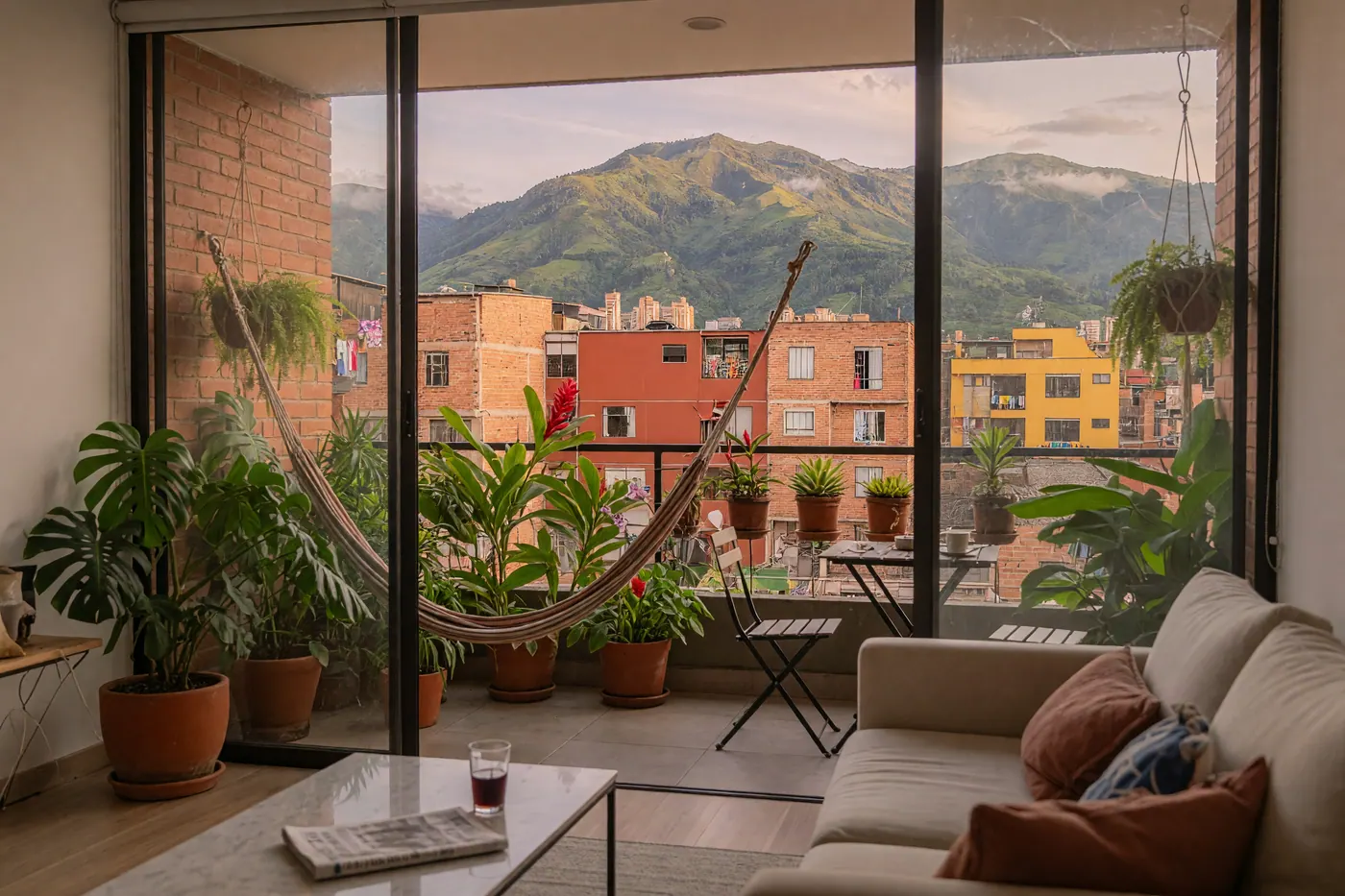 Modern Colombian apartment interior looking through glass doors to a balcony with potted plants, red-brick buildings, and green mountains.