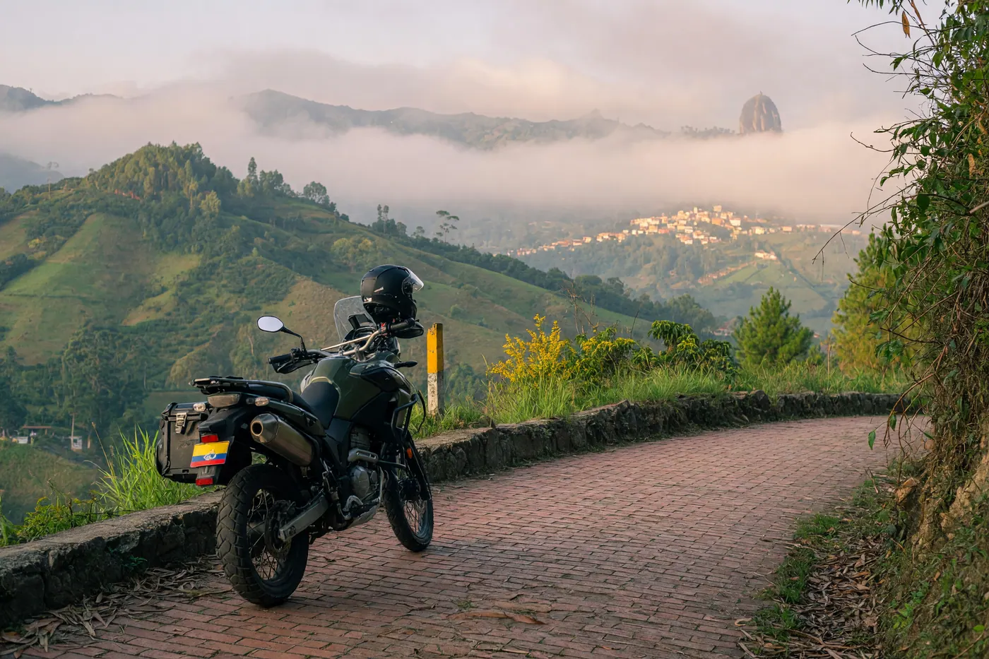 Adventure motorcycle parked on a brick Andean mountain road with a helmet on the seat and misty green hills near Guatape