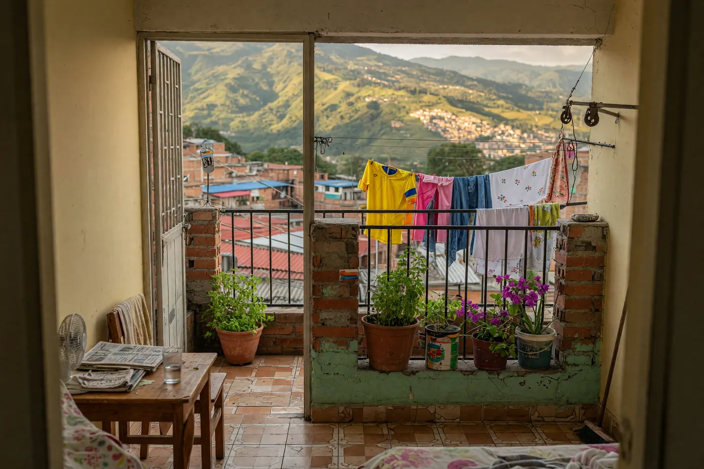 Daylight view from a furnished apartment toward a tiled balcony with plants, laundry on a rail, and hills beyond