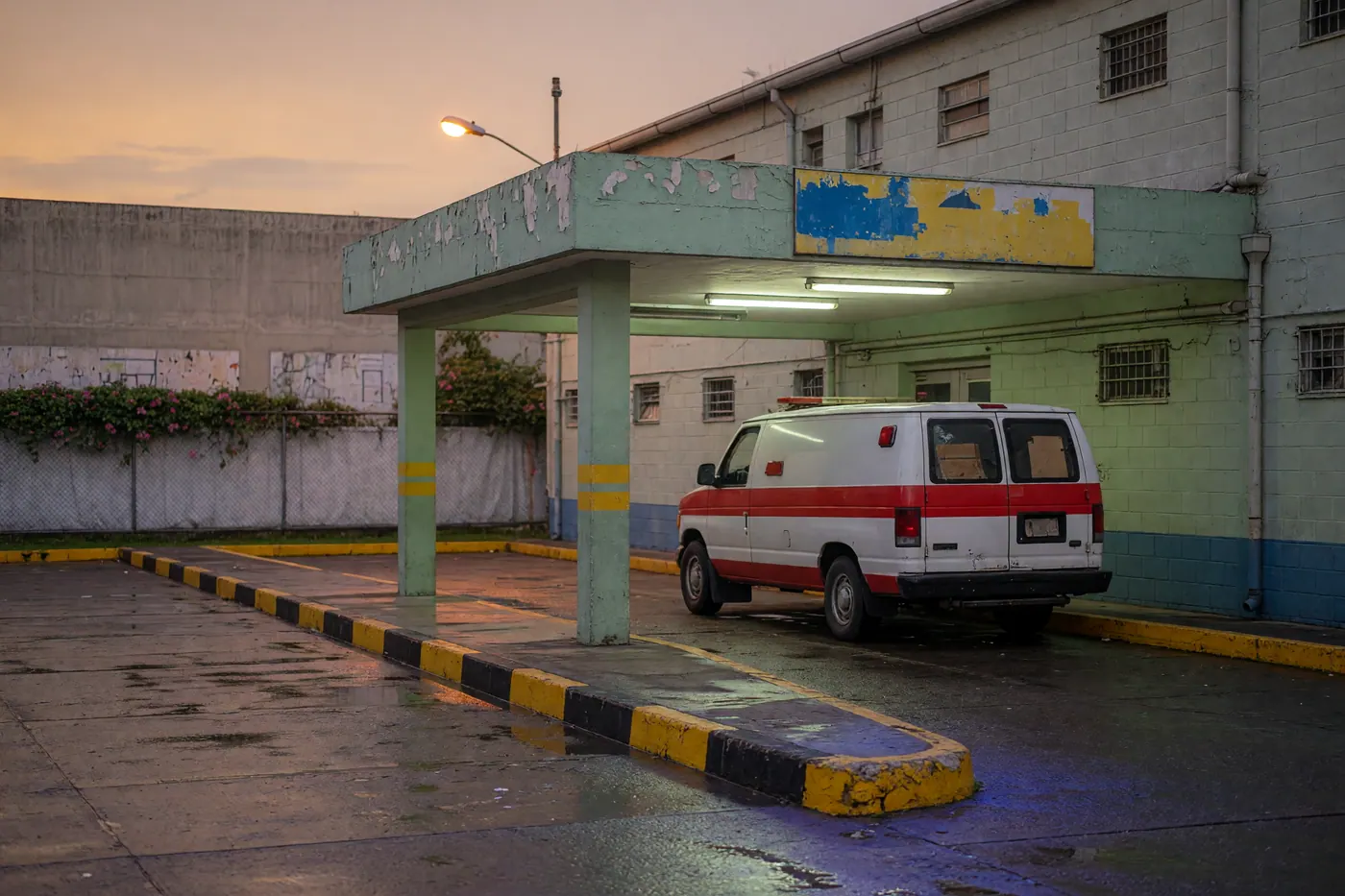 White ambulance under a fluorescent concrete emergency canopy at dusk with yellow-black curbs