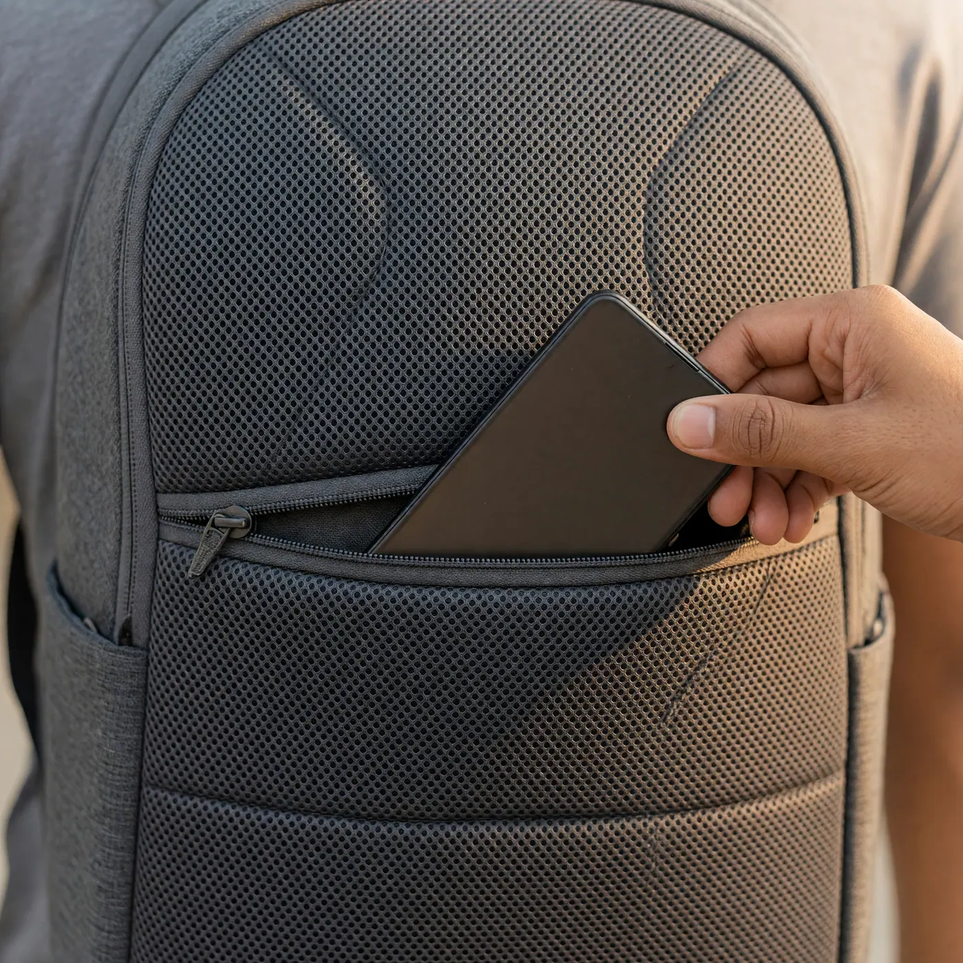 Mano deslizando un celular hacia el bolsillo oculto en el espaldar de una mochila gris — protección antirrobo en el TransMilenio