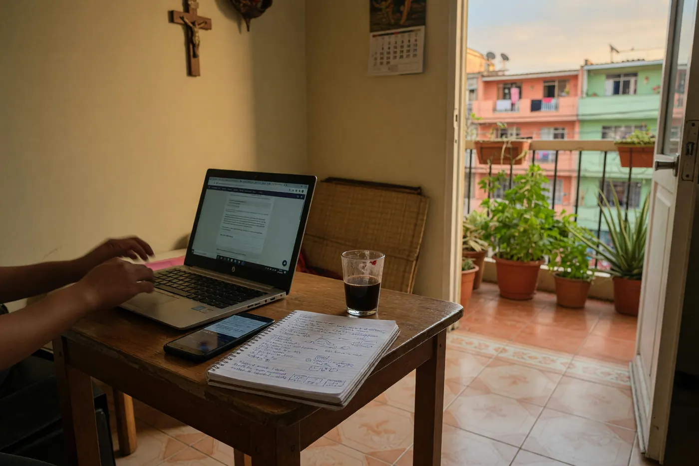 A laptop and phone on a small table in a modest Colombian apartment with a notebook and balcony plants in soft daylight