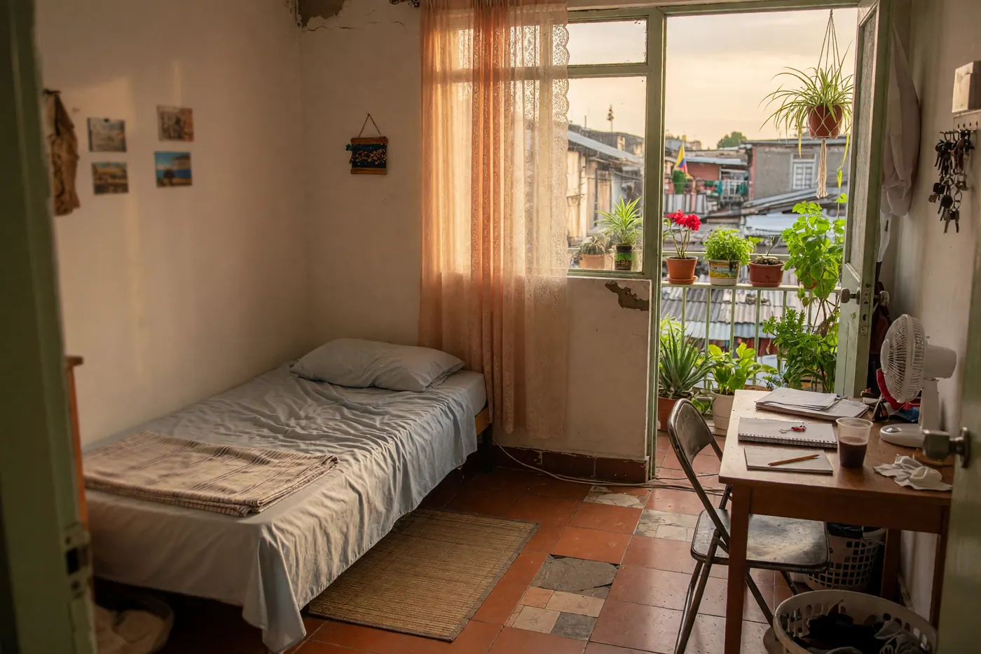 A modest, bright room in a Colombian shared apartment with a simple bed, small desk, and a window with balcony plants
