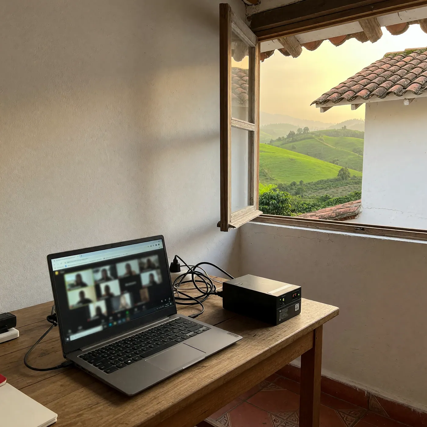 Laptop showing a video call beside a UPS on a finca desk overlooking green hills