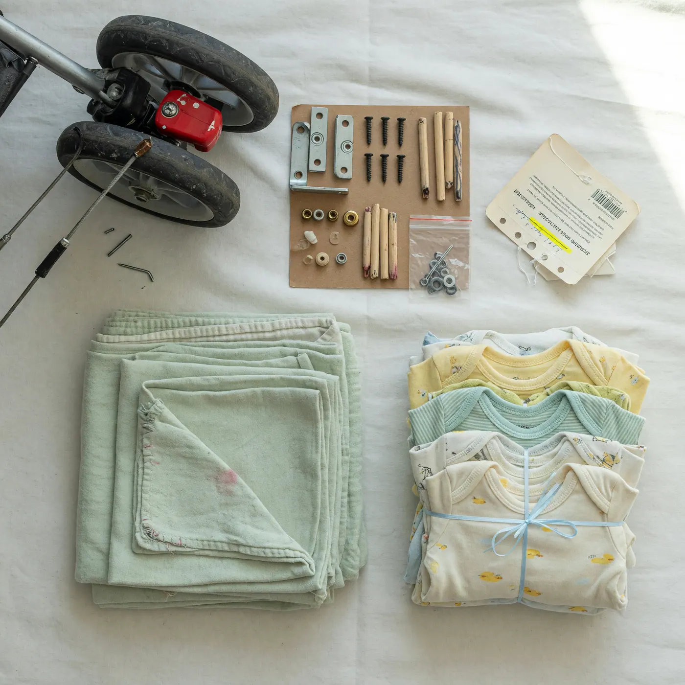 Detalle de revisión de artículos de bebé usados antes de venderlos: ruedas y freno de un coche, tornillos de una cuna, una etiqueta de modelo, tela lavada y un paquete de ropa de bebé doblada