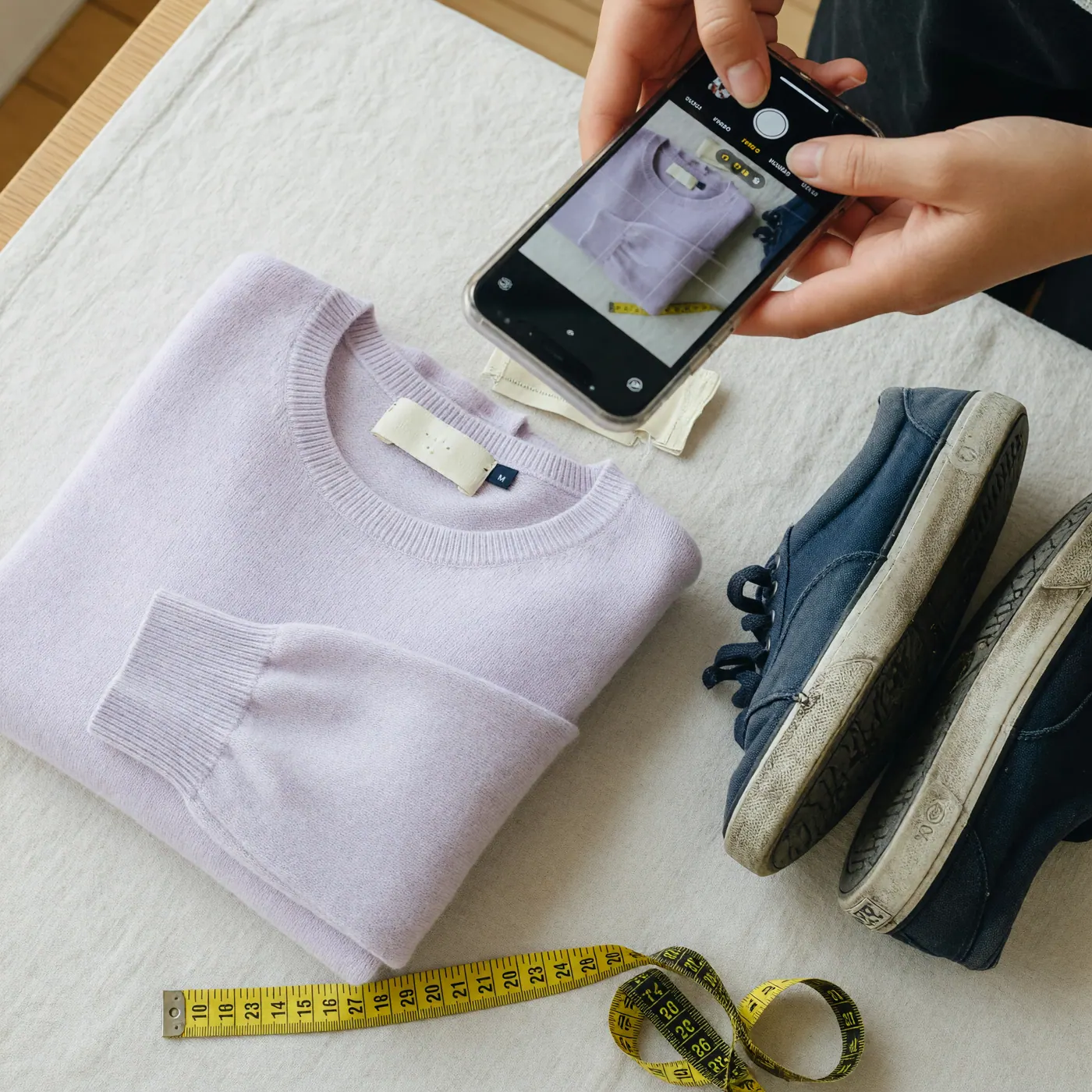 Persona fotografiando con el celular un suéter usado junto a su etiqueta de talla, unos tenis y una cinta métrica para hacer un anuncio.